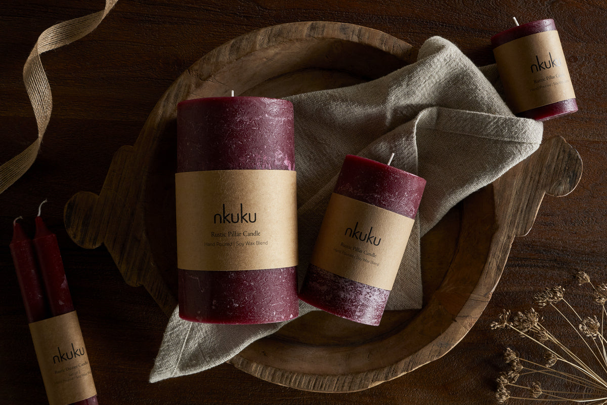 A wooden tray displays two cherry candles labeled “nkuku” and a beige cloth. Beside it, an nkuku Rustic Soy Blend Pillar Candle in Dark Cherry (Large) and two red taper candles rest on dark wood, with dried flowers nearby.