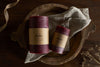 Two nkuku Rustic Soy Blend Pillar Candles in Dark Cherry (Small) sit on a beige cloth in a wooden tray, with dried flowers and ribbon on a dark wooden surface for an elegant, rustic display.