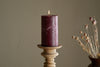 A lit nkuku Rustic Soy Blend Pillar Candle in Dark Cherry, large size, rests on a wooden holder. The background is neutral with a vase of dried flowers partly visible to the right.