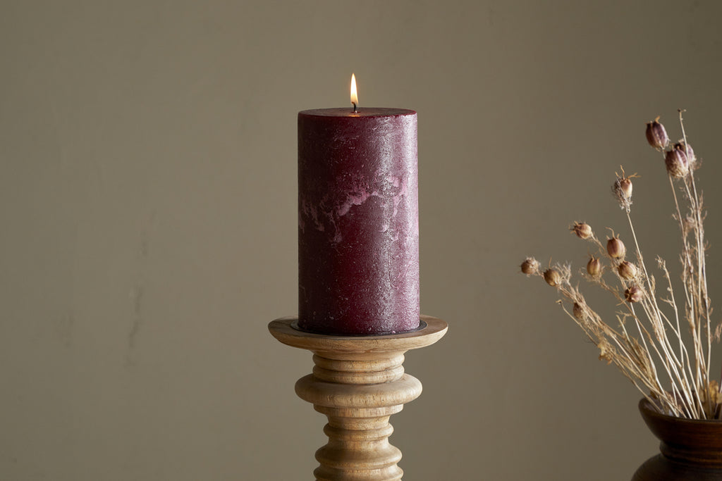 A lit nkuku Rustic Soy Blend Pillar Candle in Dark Cherry, large size, rests on a wooden holder. The background is neutral with a vase of dried flowers partly visible to the right.