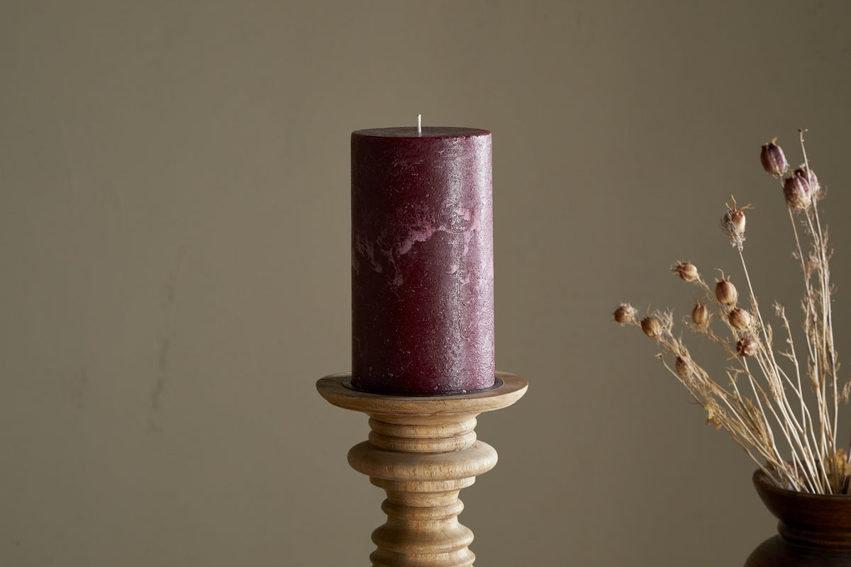 A nkuku Rustic Soy Blend Pillar Candle in Dark Cherry—large, with a textured finish—rests on a wooden holder. To the right, dried flowers in a brown vase contrast against a plain beige wall.