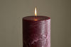 The nkuku Rustic Soy Blend Pillar Candle - Dark Cherry - Large glows softly with a steady flame, its textured finish in rich cherry hues standing out against a simple, light background.