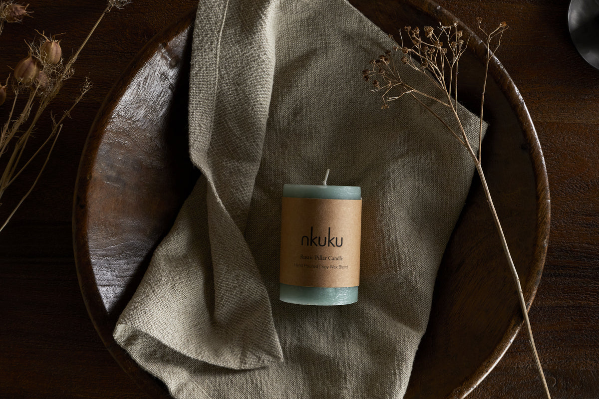 A Rustic Soy Blend Mini Pillar Candle in Sage Green by nkuku, featuring a textured finish, rests on a beige cloth in a wooden bowl with dried plants, atop a dark wooden surface.