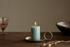 A lit nkuku Rustic Soy Blend Mini Pillar Candle in sage green with a textured finish sits on a brass holder with handle on a wooden table, accompanied by a clear glass, wooden bowl, and dried flowers against a neutral backdrop.
