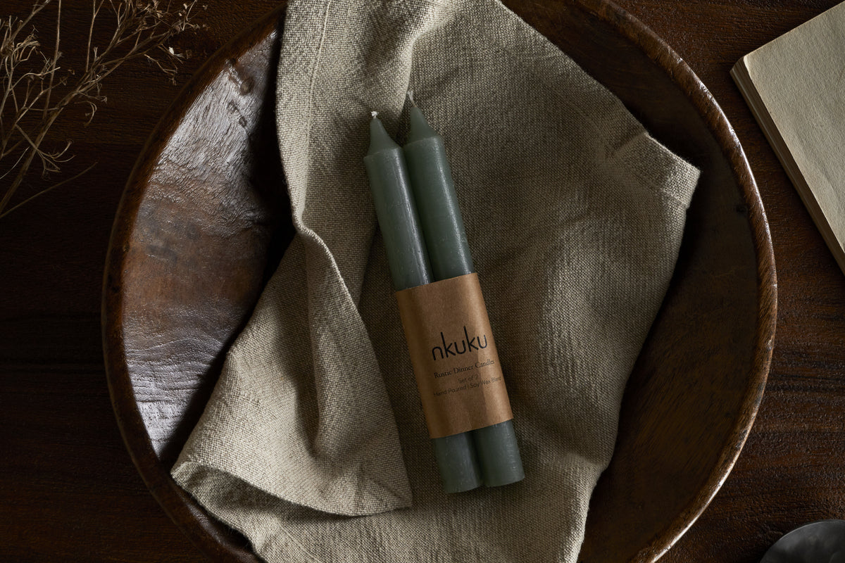 A set of two nkuku Rustic Soy Blend Dinner Candles in sage green, wrapped in a branded label, rest on beige cloth in a wooden bowl, with a dried plant and part of an open book on a dark wooden surface.