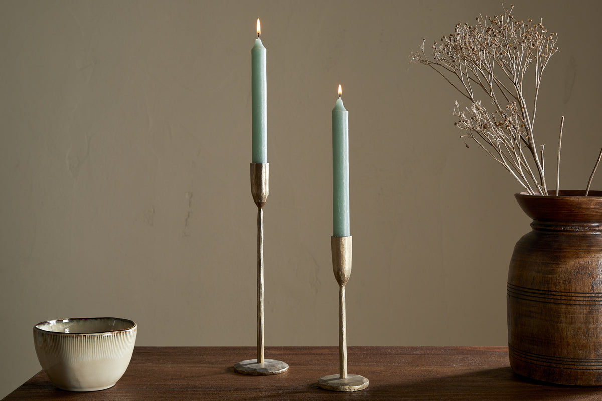 A wooden table features nkukus Rustic Soy Blend Dinner Candle - Sage Green (Set of 2) in brass holders, a ceramic bowl, and a brown vase with dried branches, all set against a beige wall.