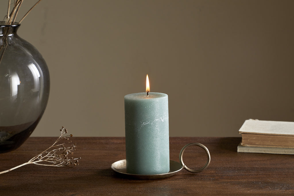 A lit nkuku Rustic Soy Blend Pillar Candle in sage green, small size, stands on a metal holder with a ring handle atop a wooden table, beside dried flowers, a dark glass vase with twigs, and a closed book. The background is plain and neutral.