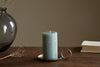 The nkuku Rustic Soy Blend Pillar Candle in sage green sits on a metal holder with a circular handle on a wooden table, next to a glass vase, dried flowers, and a light-covered book.