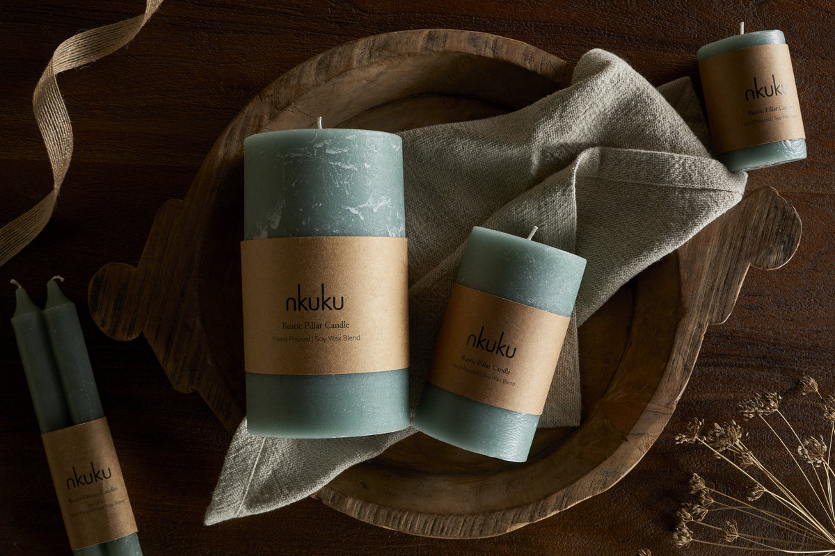 A wooden tray displays the nkuku Rustic Soy Blend Pillar Candle in large sage green, featuring a textured finish and brown label, set on a beige cloth napkin. More sage candles and dried flowers sit nearby on a dark wood surface.