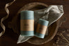 Two nkuku Rustic Soy Blend Pillar Candles in sage green with a textured finish rest on a grey cloth inside a wooden bowl, set atop a dark surface, accented by ribbon and dried flowers. Each candle features the brown paper label.