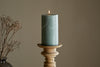 The nkuku Rustic Soy Blend Pillar Candle in Sage Green (Large) with a textured finish sits on a wooden holder, accompanied by dried flowers in a black vase to the left, all set against a plain beige background.