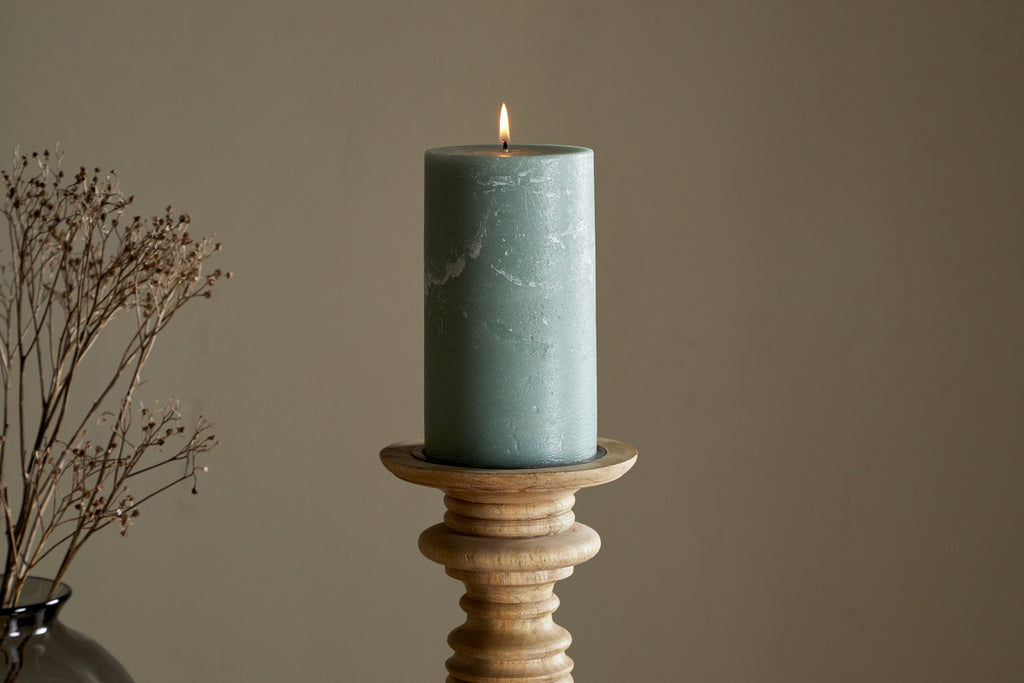 The nkuku Rustic Soy Blend Pillar Candle in Sage Green (Large) with a textured finish sits on a wooden holder, accompanied by dried flowers in a black vase to the left, all set against a plain beige background.