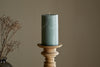 A Rustic Soy Blend Pillar Candle in Sage Green by nkuku sits on a wooden holder, with dried branches in a vase partially visible on the left, all set against a neutral beige background.