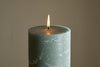 A close-up of the nkuku Rustic Soy Blend Pillar Candle in Sage Green (Large), featuring a textured surface and burning with a small flame against a plain beige background.