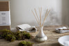 A nkuku Sea Salt & Moss Reed Diffuser with wooden sticks rests on a rustic table, surrounded by green moss, a linen cloth, its box, a wooden spoon filled with salt, and a white ceramic bowl for an inviting fragrance display.