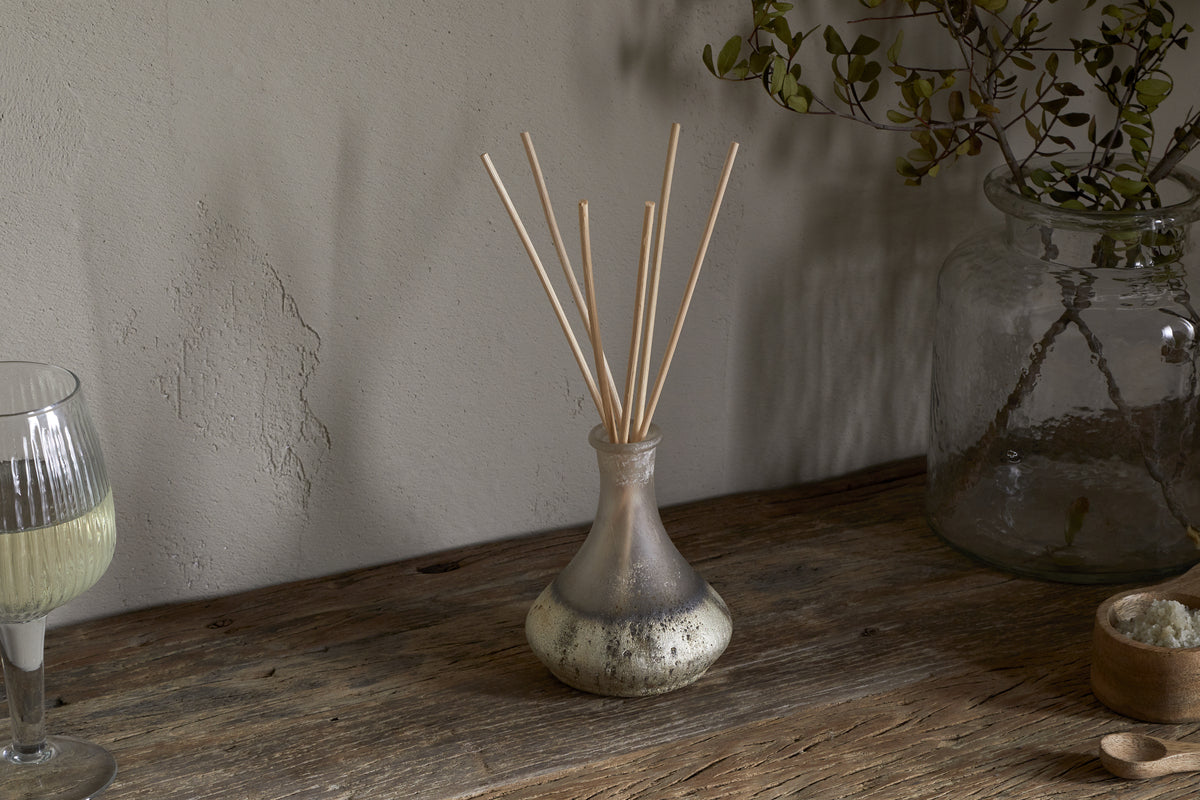 The nkuku Sea Salt & Moss Reed Diffuser adds a bespoke scent to a rustic wooden table, alongside a partial glass of white wine, a large jar with foliage, and a small bowl of salts.