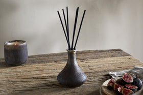 A nkuku Cedarwood & Fig Reed Diffuser with black reeds stands on a rustic wooden table, accompanied by a dark candle and fresh figs, infusing the air with a cozy natural cedarwood scent.