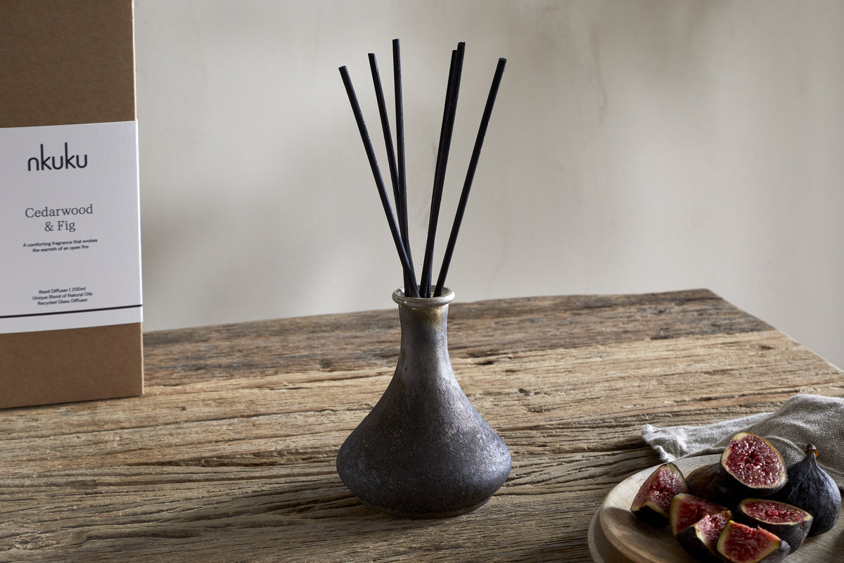 A nkuku Cedarwood & Fig Reed Diffuser with black reeds sits on a wooden table beside sliced figs and its box, filling the room with the warm scent of cedarwood.