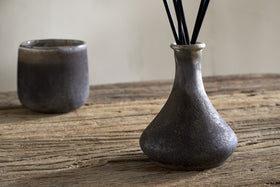 A nkuku Cedarwood & Fig Reed Diffuser with slender reeds and a matching cup rest on a rustic wooden table, set against a neutral backdrop.
