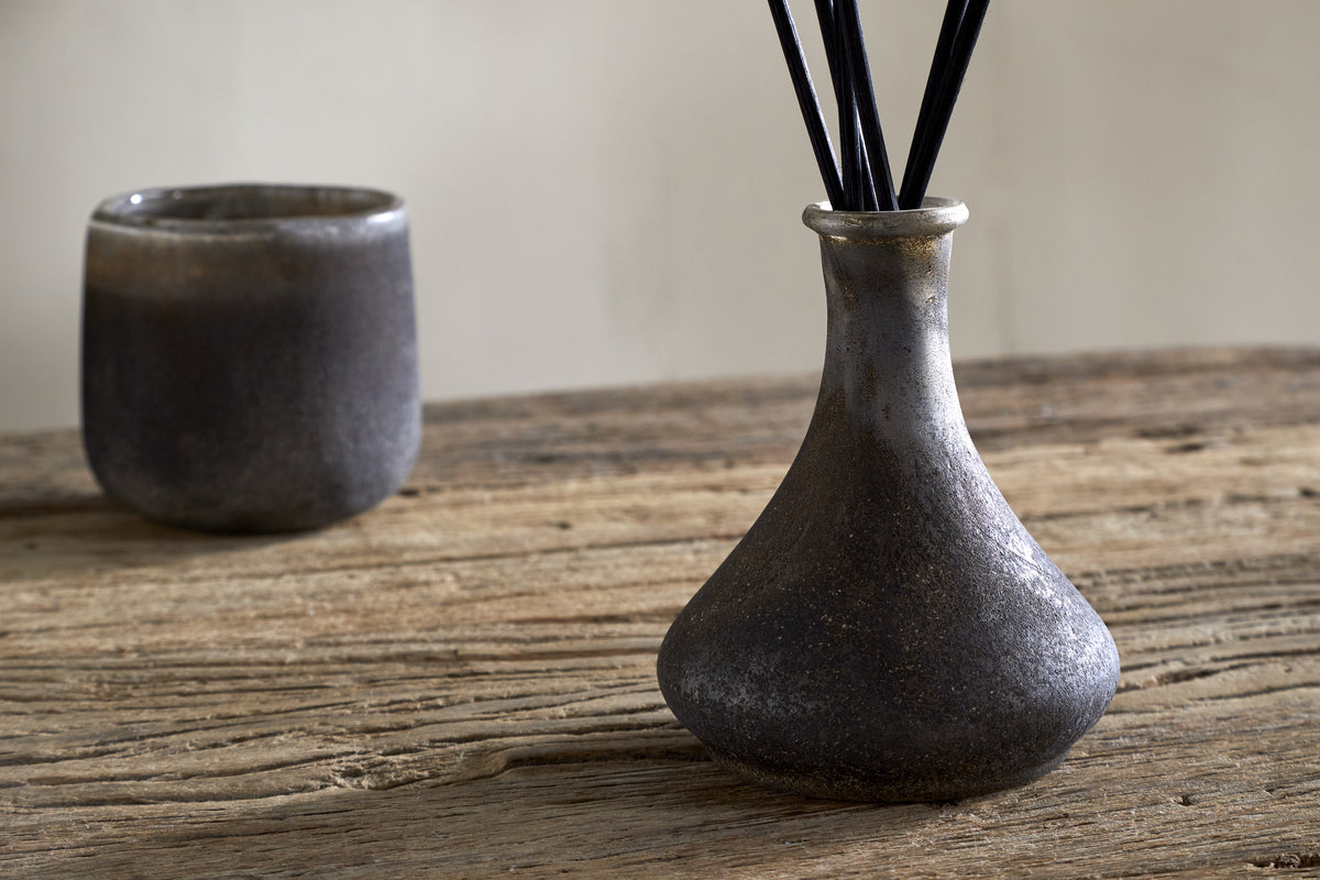 A nkuku Cedarwood & Fig Reed Diffuser with slender reeds and a matching cup rest on a rustic wooden table, set against a neutral backdrop.