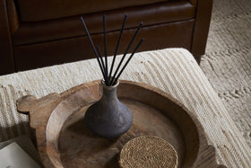 The nkuku Cedarwood & Fig Reed Diffuser, with black reeds in a dark ceramic vase, sits on a rustic wooden tray beside a woven coaster atop a textured ottoman, filling the air with cedarwood aroma.