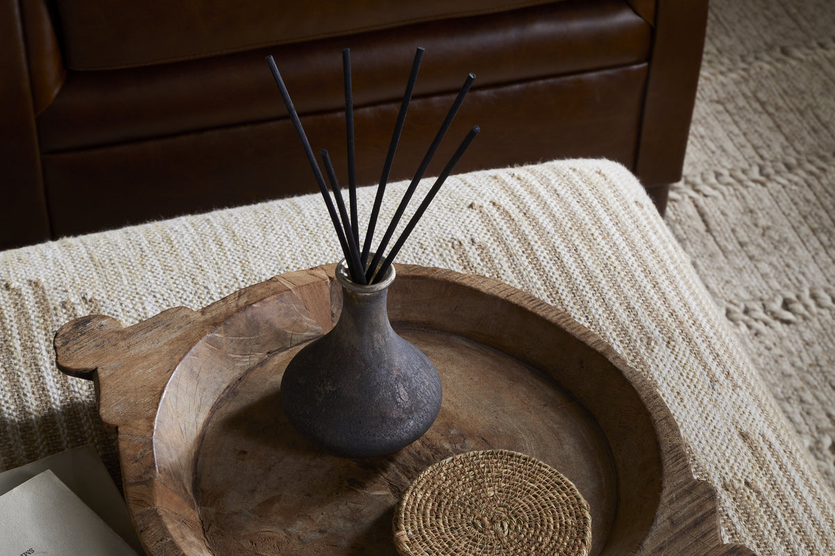 The nkuku Cedarwood & Fig Reed Diffuser, with black reeds in a dark ceramic vase, sits on a rustic wooden tray beside a woven coaster atop a textured ottoman, filling the air with cedarwood aroma.