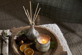 The nkuku Mandarin & Clove 1 Wick Scented Candle sits with festive decor on a rustic wooden tray, filling the room with spiced mandarin fragrance atop a textured rug and cushioned stool.