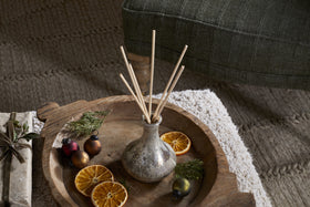A Mandarin & Clove 1 Wick Scented Candle by nkuku sits on a rustic wooden tray with dried orange slices and ornaments, displayed on a textured rug near a green upholstered chair.