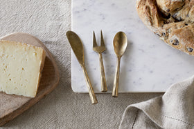 A block of cheese, bread, a beige napkin, and the nkuku Roda Serving Set – Antique Brass (Set of 3) on marble and linen create an elegant display.