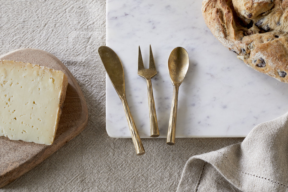 A block of cheese, bread, a beige napkin, and the nkuku Roda Serving Set – Antique Brass (Set of 3) on marble and linen create an elegant display.