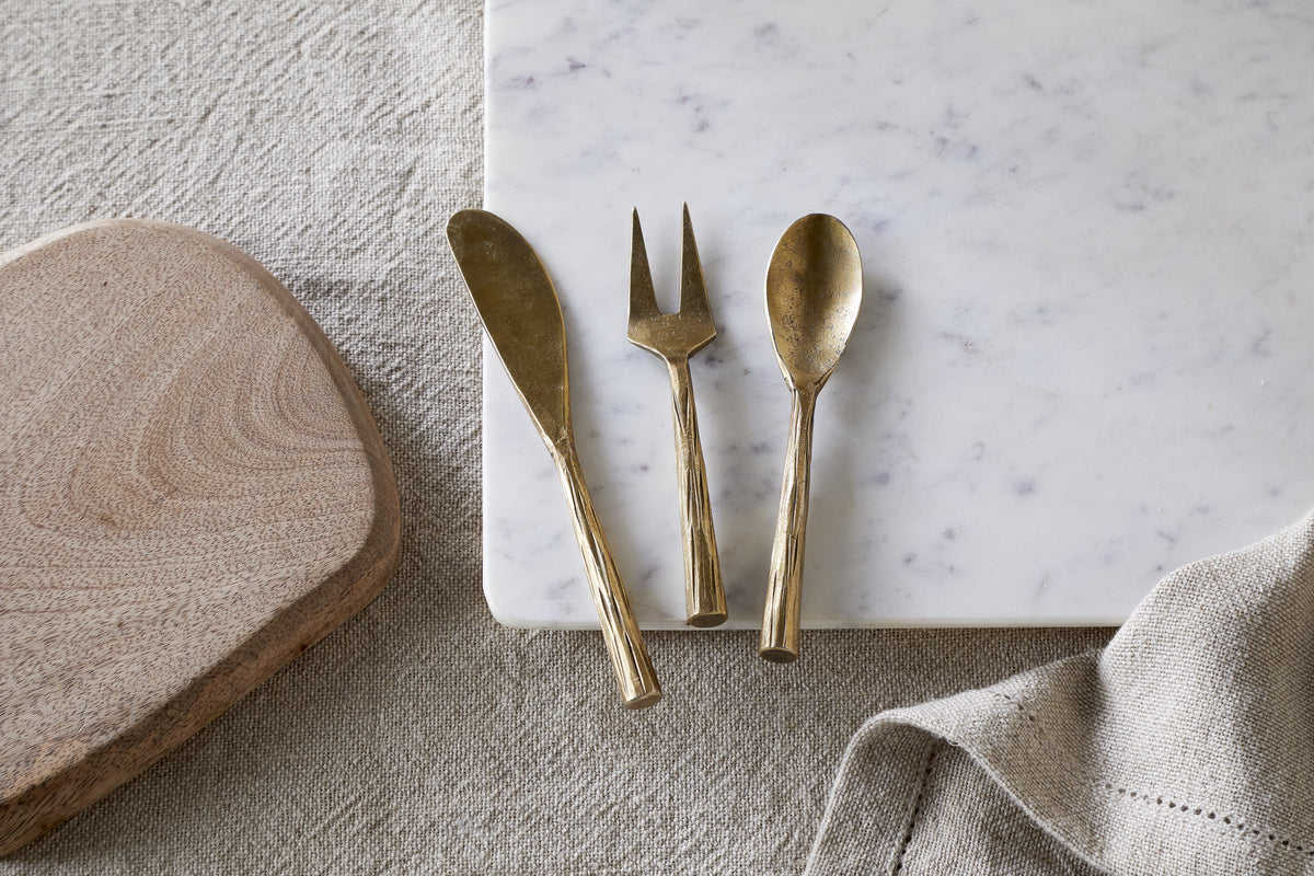 The nkuku Roda Serving Set - Antique Brass (set of 3) is displayed on a white marble slab next to a wooden cutting board and a textured beige cloth, all set atop a fabric surface.