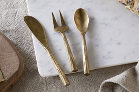 The nkuku Roda Serving Set – Antique Brass (Set of 3), featuring a spreader, fork, and spoon, rests on white marble beside bread atop beige fabric.