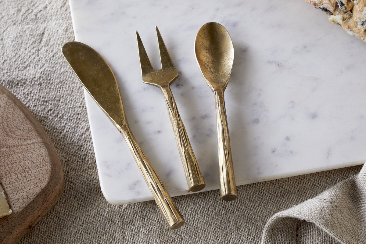 The nkuku Roda Serving Set – Antique Brass (Set of 3), featuring a spreader, fork, and spoon, rests on white marble beside bread atop beige fabric.