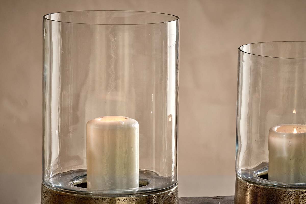 Two white pillar candles glow in nkuku Rajura Hurricane Lanterns—clear cylindrical holders with metallic bases—casting ambient light against a beige backdrop. Set side by side, one lantern is mostly out of frame to the right.