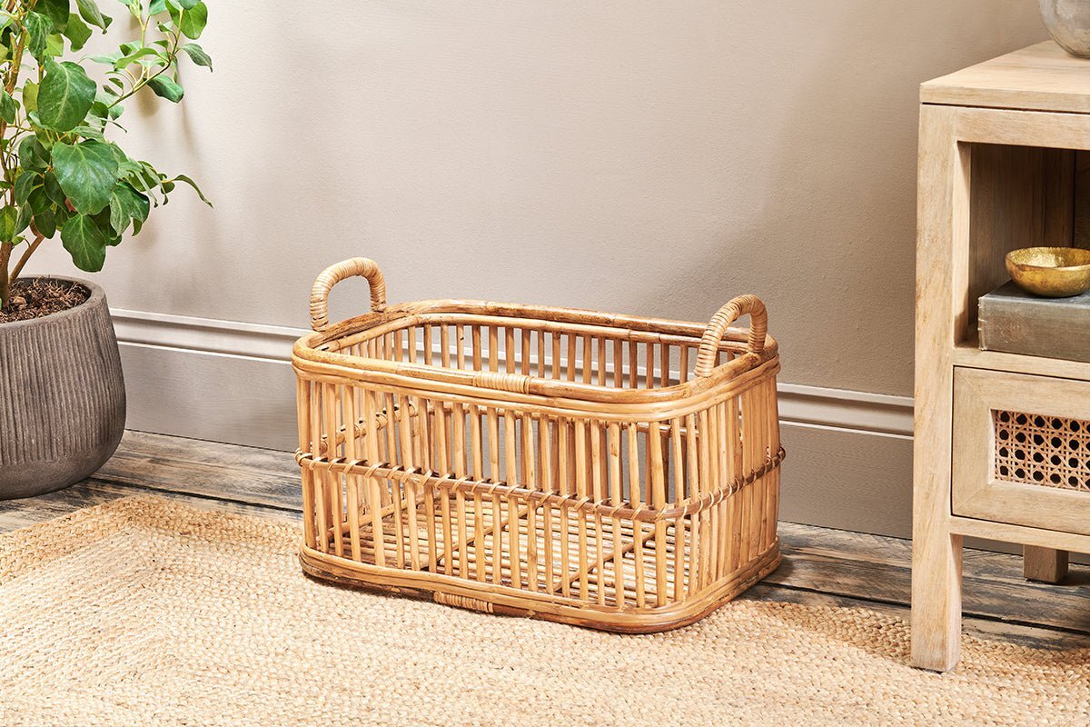 The nkuku Rammi Rattan Laundry Basket, featuring two handles and an open weave design, sits empty on a woven rug beside a light wood table and a plant in a gray pot, adding to the cozy, natural atmosphere.