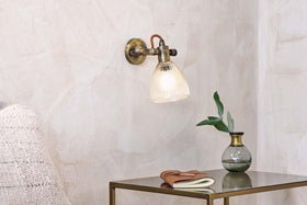 The nkuku Rarni Recycled Glass Single Wall Light casts a warm glow above a small metallic table, which holds a green glass vase with leafy branch, a closed book, and a folded brown leather item for a cozy minimalist look.