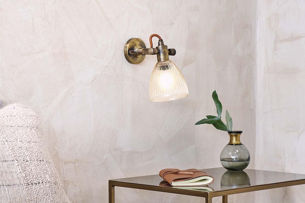 The nkuku Rarni Recycled Glass Single Wall Light casts a warm glow above a small metallic table, which holds a green glass vase with leafy branch, a closed book, and a folded brown leather item for a cozy minimalist look.
