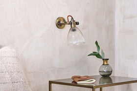 The nkuku Rarni Recycled Glass Single Wall Light is mounted on a textured beige wall above a metal side table with a green-leaf vase, closed notebook, and brown eyeglasses. The edge of a textured chair appears on the left.