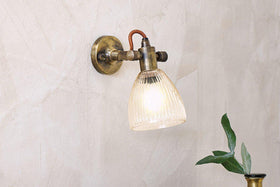 The nkuku Rarni Recycled Glass Single Wall Light is mounted on a textured beige wall, glowing warmly. Below the sconce, a vase filled with green leafy branches is partially visible.