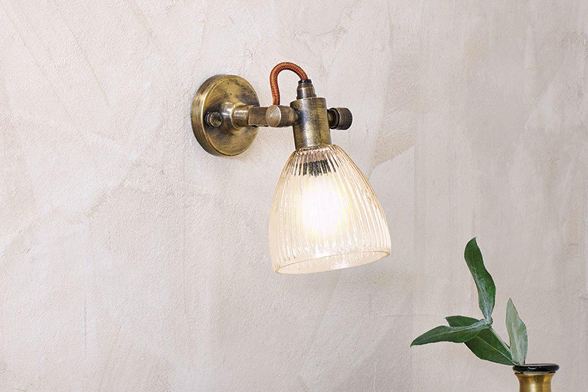 The nkuku Rarni Recycled Glass Single Wall Light is mounted on a textured beige wall, glowing warmly. Below the sconce, a vase filled with green leafy branches is partially visible.