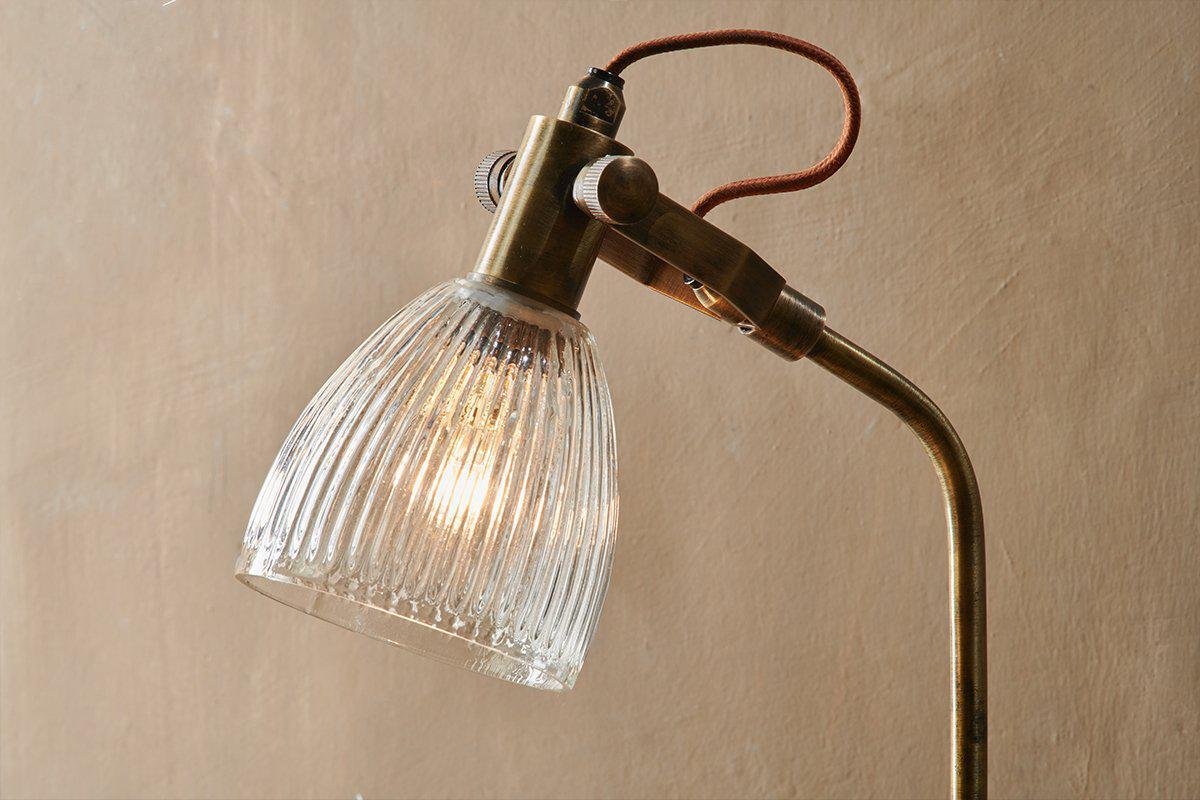 A close-up of the nkuku Rarni Recycled Glass Table Lamp in antique brass, featuring a ribbed clear glass shade, curved neck, exposed brown fabric cord, and warm light against a beige textured wall.