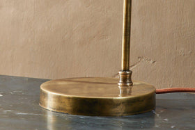 Close-up of the nkuku Rarni Recycled Glass Table Lamp in Antique Brass: a cylindrical stem and ribbed glass shade rest on a dark surface, with a brown fabric power cord extending toward a tan wall.