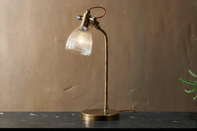 The nkuku Rarni Recycled Glass Table Lamp in Antique Brass, featuring a ribbed glass shade and curved arm, stands on dark marble. Its reddish-brown cord loops behind it against a plain brown wall, casting a warm light.