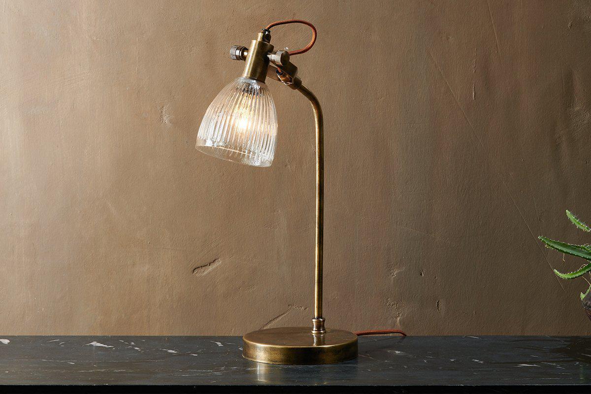 The nkuku Rarni Recycled Glass Table Lamp in Antique Brass, featuring a ribbed glass shade and curved arm, stands on dark marble. Its reddish-brown cord loops behind it against a plain brown wall, casting a warm light.