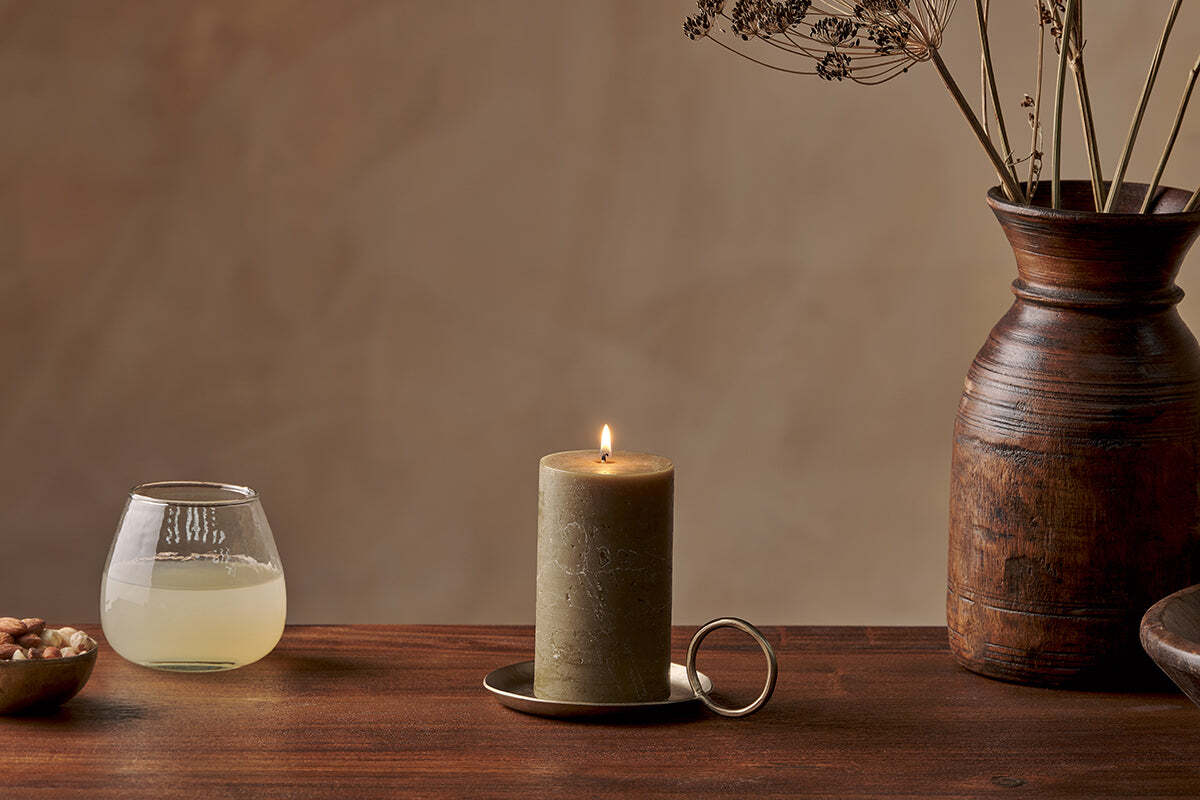 A lit Nkuku Rustic Soy Blend Pillar Candle in Olive Green (Small, textured finish) sits on a brass tray atop a wooden table, next to a clear glass of pale liquid, a bowl of nuts, and a tall wooden vase with dried flowers against a beige background.