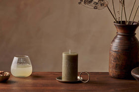 The Nkuku Rustic Soy Blend Pillar Candle in olive green sits on a brass holder atop a wooden table, flanked by a glass of pale yellow liquid and nuts on the left and a brown ceramic vase with dried flowers on the right.