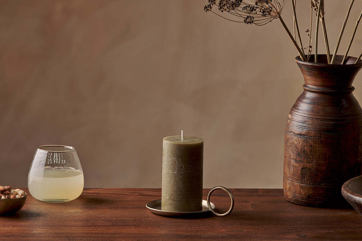 The Nkuku Rustic Soy Blend Pillar Candle in olive green sits on a brass holder atop a wooden table, flanked by a glass of pale yellow liquid and nuts on the left and a brown ceramic vase with dried flowers on the right.