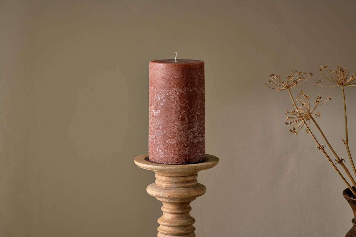 The nkuku Rustic Soy Blend Pillar Candle - Rust - Large, featuring a textured finish, is displayed on a wooden candlestick holder against a beige backdrop, with dried plant stems to the right for a natural accent.