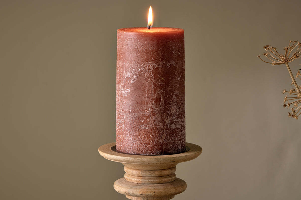 A lit nkuku Rustic Soy Blend Pillar Candle - Rust - Large with a textured finish sits on a round wooden holder against a plain beige background. A dry plant is faintly visible to the right.