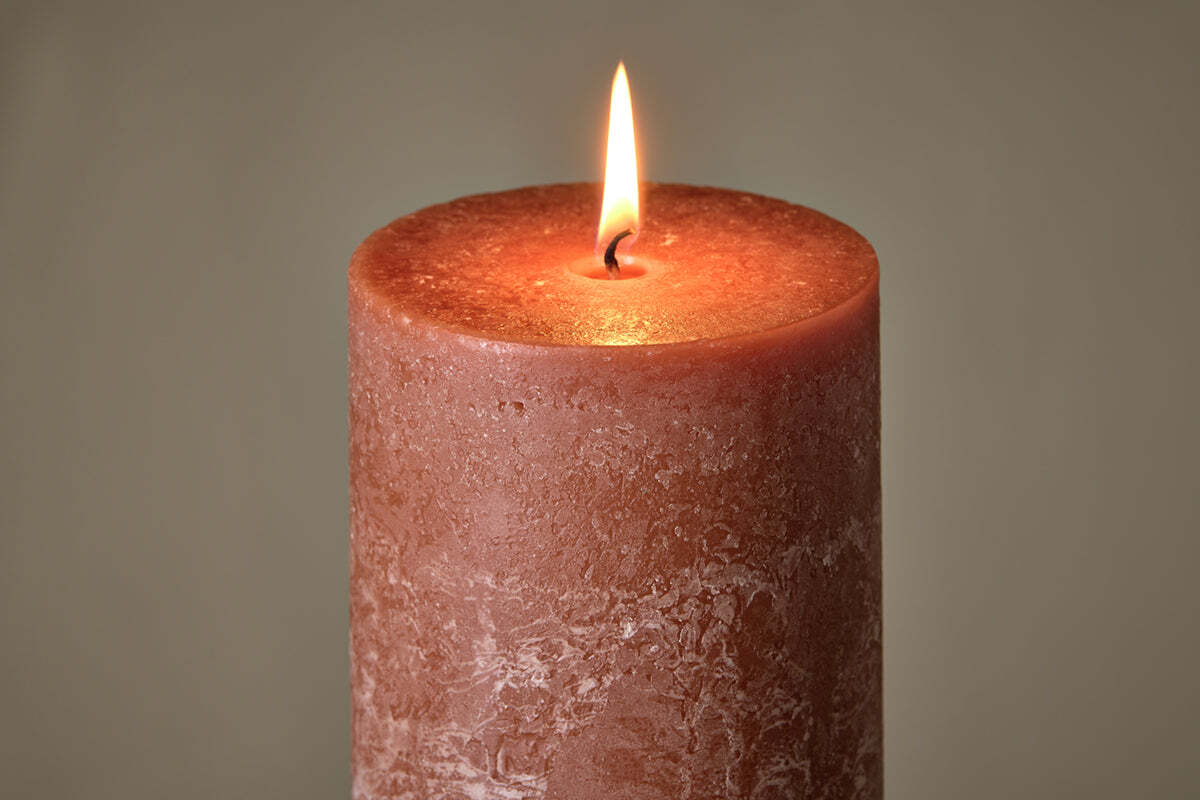 A close-up of the nkuku Rustic Soy Blend Pillar Candle in Rust (Large) showcases its textured reddish-brown wax, unique imperfections, and a warm flame glowing at the top against a softly blurred background.